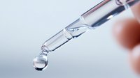 Clear liquid drop falling from glass pipette or dropper against blurred light background, macro photography showing surface tension and transparency.