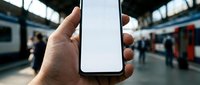 Hand holding blank smartphone screen at busy train station platform with commuters and trains in blurred background for mobile app mockup presentations.