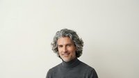 Middle-aged man with gray wavy hair and light skin tone wearing dark turtleneck sweater, smiling at camera against neutral background for portrait use.