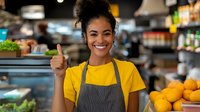 Young African American female retail worker in yellow t-shirt and gray apron showing thumbs up while standing near fresh produce display in grocery store, expressing positivity.