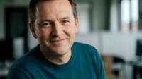 Middle-aged Caucasian businessman in teal sweater smiling confidently in modern office environment for corporate communications and professional marketing.