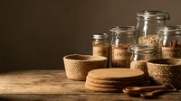 Glass jars with labeled lids containing dry goods like oats and lentils, arranged with woven baskets and wooden spoons on rustic table for pantry organization and storage.
