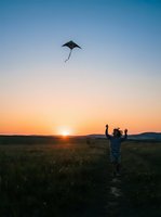 Person flying kite in open field at sunset, wearing casual clothing with arms raised, golden hour lighting over rural landscape with mountains in distance.