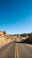Straight asphalt highway with yellow center lines stretching through arid desert landscape with rock formations under clear blue sky, suitable for travel and road trip concepts.