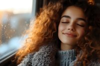 Young woman with curly hair relaxing by window in cozy sweater during golden hour sunlight for wellness and self-care content.