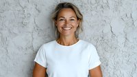 Confident mature blonde woman in white t-shirt smiling against textured gray wall background. Natural daylight portrait showcasing genuine happiness and positive attitude.