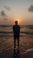 Silhouette of young man standing alone on beach at sunset, watching waves roll in. Dramatic orange sky and clouds reflect in wet sand, creating peaceful seaside meditation scene.