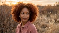Young African American woman with curly afro hair smiling outdoors in golden hour light wearing casual shirt in natural field setting for lifestyle photography.