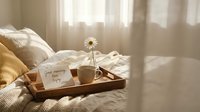 Cozy morning bedroom scene with breakfast tray on unmade bed featuring coffee cup, flower vase and good morning card in warm sunlight streaming through curtains.