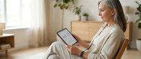 Senior Caucasian woman in beige loungewear using tablet computer while relaxing in comfortable wooden chair at bright modern home interior with plants.