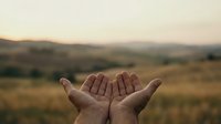 Open hands reaching toward golden sunset sky over rolling countryside landscape symbolizing hope, prayer, and spiritual connection with nature.
