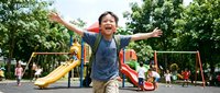Happy Asian boy with arms spread wide enjoying outdoor playground with colorful slides and swings in sunny park setting for family recreation concepts.