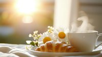 Fresh baked croissant and steaming cup of coffee with chamomile flowers on wooden table in morning sunlight, creating cozy breakfast atmosphere.