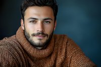 Confident young Mediterranean man with beard wearing brown turtleneck sweater against dark blue background, looking at camera with gentle smile.