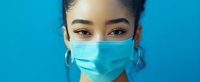 Young Asian woman wearing protective medical face mask looking at camera with confident eyes against blue background, healthcare concept.
