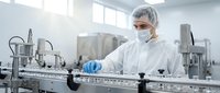 Female pharmaceutical technician in protective suit and mask operating sterile production line equipment in modern clean room manufacturing facility.