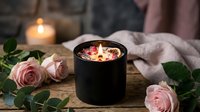 Luxury scented candle with dried flowers in black container surrounded by pink roses on wooden surface creating romantic spa atmosphere for relaxation.