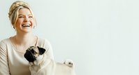 Young Caucasian woman with blonde hair in updo hairstyle holding and playing with cute pug dog. Both smiling happily against white background for pet care advertising.