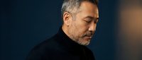 Middle-aged Caucasian man with gray beard in black turtleneck sweater looking down pensively against dark background with warm lighting for portraits.