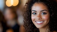 Young African American woman with natural curly hair and bright white smile against blurred bokeh background, radiating warmth and confidence in casual setting.