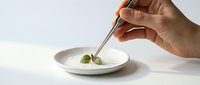 Chef's hand using tweezers to plate a fresh basil leaf on a white ceramic dish, showcasing fine dining precision and culinary artistry.