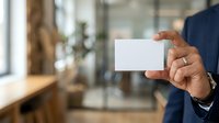 Businessman in navy suit holding blank white business card in modern office environment with natural light and glass partitions.