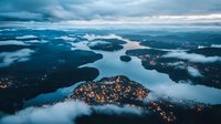 Aerial view of illuminated coastal town nestled between fjords and mountains at dusk, with low-lying clouds creating mystical atmosphere over calm waters.