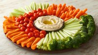 Fresh vegetable platter with hummus dip arranged in circular pattern featuring carrots, cherry tomatoes, celery, broccoli on marble surface for party appetizer.