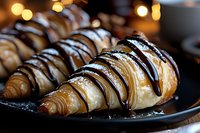 Fresh baked croissants drizzled with dark chocolate and white chocolate sauce on black plate, close up view with soft bokeh lights in background.