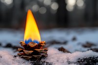 Burning pinecone candle glowing with bright orange flame on snowy ground against blurred bokeh background creates cozy winter atmosphere.