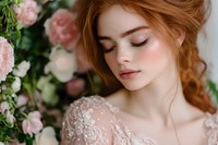Young Caucasian bride with red hair in lace wedding dress among pink roses, eyes closed in peaceful moment. Romantic bridal portrait for wedding photography.