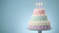 Three-tier birthday cake with pastel pink, mint green and lavender frosting rosettes, lit candles on blue background for celebration designs.