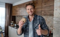 Cheerful Caucasian man in plaid shirt showing thumbs up while holding cookie in modern kitchen with wooden cabinetry.