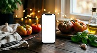 Smartphone with blank white screen mockup on rustic kitchen table surrounded by fresh tomatoes, herbs and cooking ingredients for food app design.