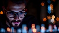 Young man with beard and glasses working on computer at night with blue screen light reflection and colorful bokeh background.