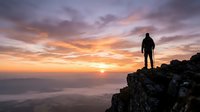 Silhouetted hiker standing on rocky mountain peak at sunset, watching golden sun descend over misty valley landscape. Adventure and exploration concept.