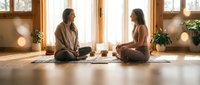 Two women practicing meditation together in bright wooden studio with plants and natural light streaming through windows.