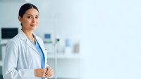 Young Hispanic female doctor in white coat with stethoscope standing confidently in modern medical office, professional healthcare worker portrait.