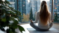 Young woman with long hair in grey activewear practicing meditation in lotus pose on yoga mat near panoramic window with urban city view and morning light.