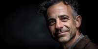 Middle-aged Caucasian man with curly gray hair and beard smiling warmly at camera against dark background for professional headshot photography.