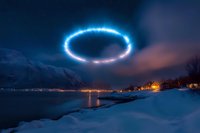 Glowing blue ring hovers over snowy winter landscape with fjord and mountain village at night, mysterious lights reflecting on calm water surface.