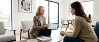 Middle-aged woman with long blonde hair and light skin tone, wearing a beige blazer, sits on a modern chair holding a laptop while consulting with a younger woman with long dark hair and olive skin tone wearing a tan sweater. Both are engaged in professional discussion in a bright, contemporary office space with minimalist decor, large windows, and modern furniture. Suitable for business, consulting, mentoring, and corporate communication concepts.