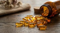 Omega-3 fish oil capsules spilling from amber glass bottle on rustic wooden surface with dried herbs for natural health and wellness supplements.