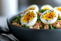 Healthy breakfast bowl with soft boiled eggs, tuna, greens and black sesame seeds in dark ceramic dish for nutritious meal preparation and diet recipes.