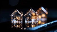 Miniature glass houses with warm glowing lights creating magical holiday atmosphere, reflected on dark glossy surface with bokeh effect in background.