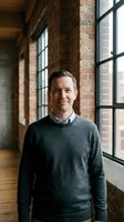 Professional Caucasian businessman in gray sweater standing confidently in modern brick loft office with large industrial windows and natural lighting.