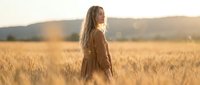 Young woman with long blonde hair standing in golden wheat field during sunset with warm light and mountains in background for lifestyle photography.