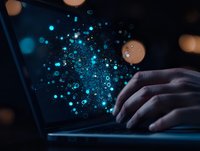 Glowing blue digital particles emerge from laptop screen while person types in dark room, creating magical technology visualization effect.