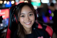 Young Asian woman gamer wearing headphones smiles at camera in gaming chair with colorful bokeh lights in background, expressing joy and confidence.