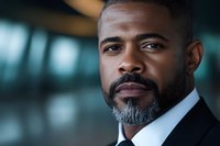 Confident African American businessman with salt and pepper beard wearing black suit, looking ahead with determined expression against blurred office background.